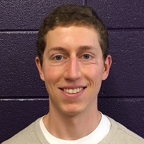 PhD student Michael Rosenberg smiles in front of a purple wall wearing a tan sweater.