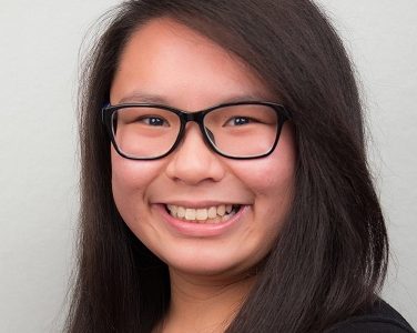 Larissa, an undergraduate researcher, stands in front of a white wall wearing black glasses and a black shirt.
