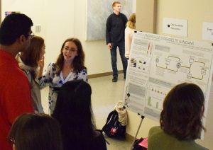 Claire Mitchell, and undergraduate student in the Steele Lab, stands in front of her poster at Mary Gates Hall during the undergraduate research symposium. Claire is wearing a white and blue floral blouse. She is in the middle of describing her research project to four community members who have taken an interest in her research.