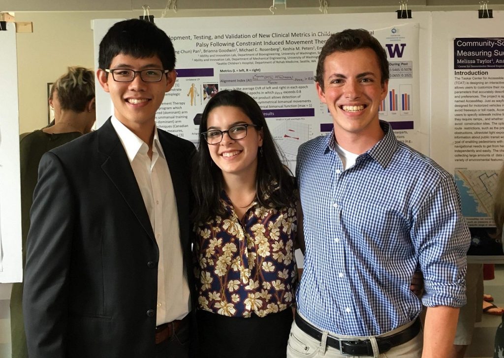 Three undergraduates, in their early twenties, stand arm in arm as they smile for the photo. They are dressed in business casual attire and behind them hangs a series of scientific posters on biomechanics.