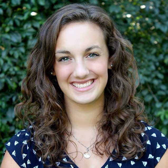 Megan smiles at the camera in front of a green leaf background. She has brown, curly hair (shoulder length) and wears a blue and white polka dot blouse.