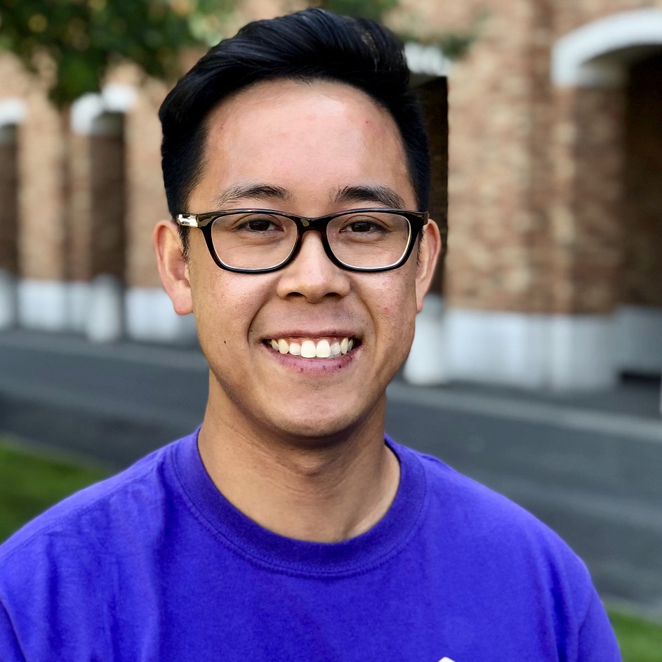 Brandon smiles at camera on the University of Washington campus in a bright purple HuskyADAPT t-shirt.