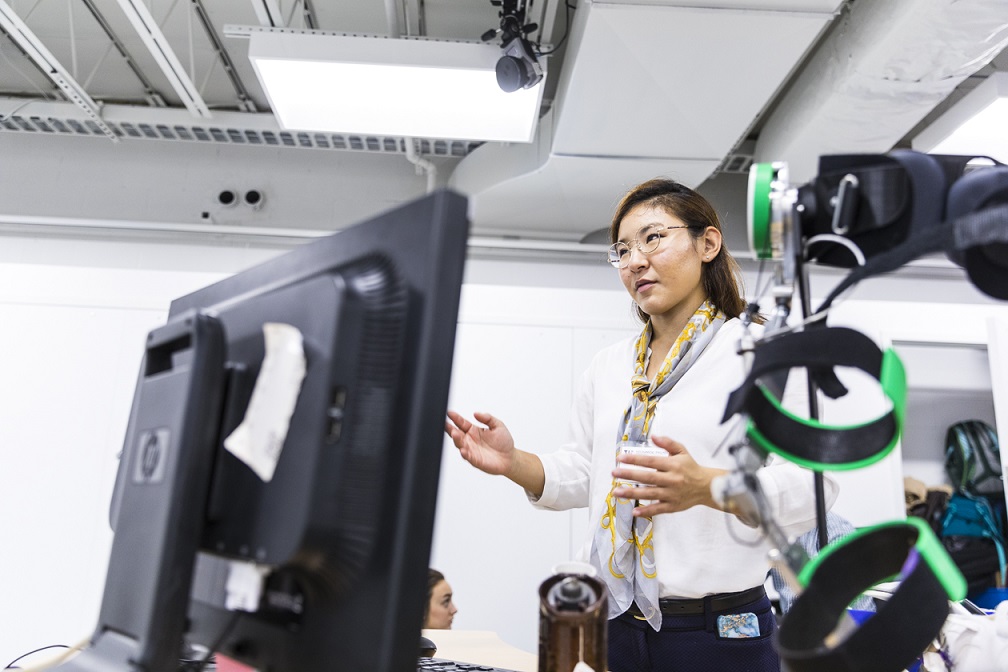 Momona discusses research in front of a computer in a large lab with white walls. There is a green and black pediatric exoskeleton in the foreground.