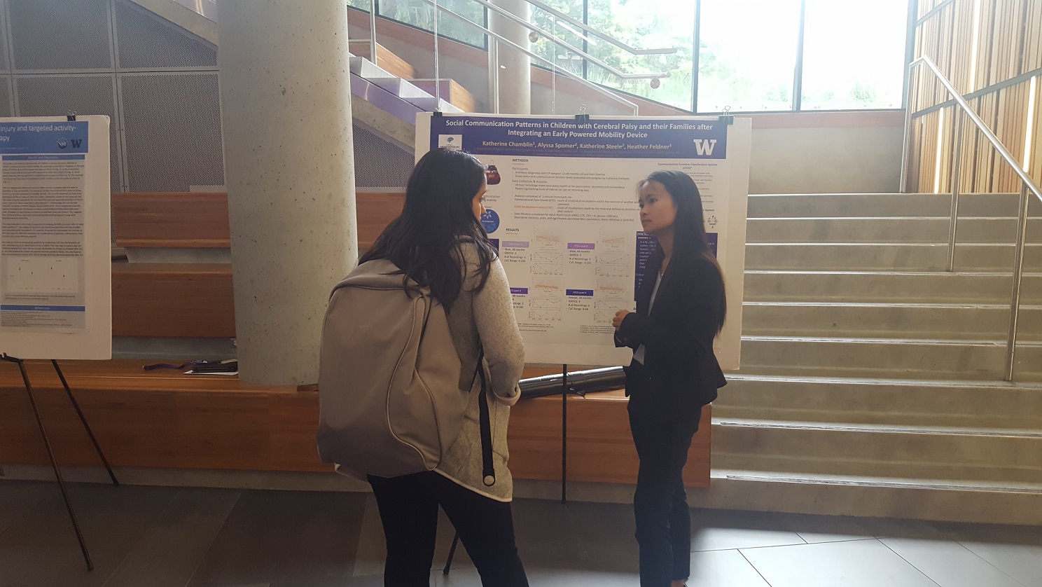 Sun shinning down on young woman in business attire talking to another woman in front of a white and purple poster board