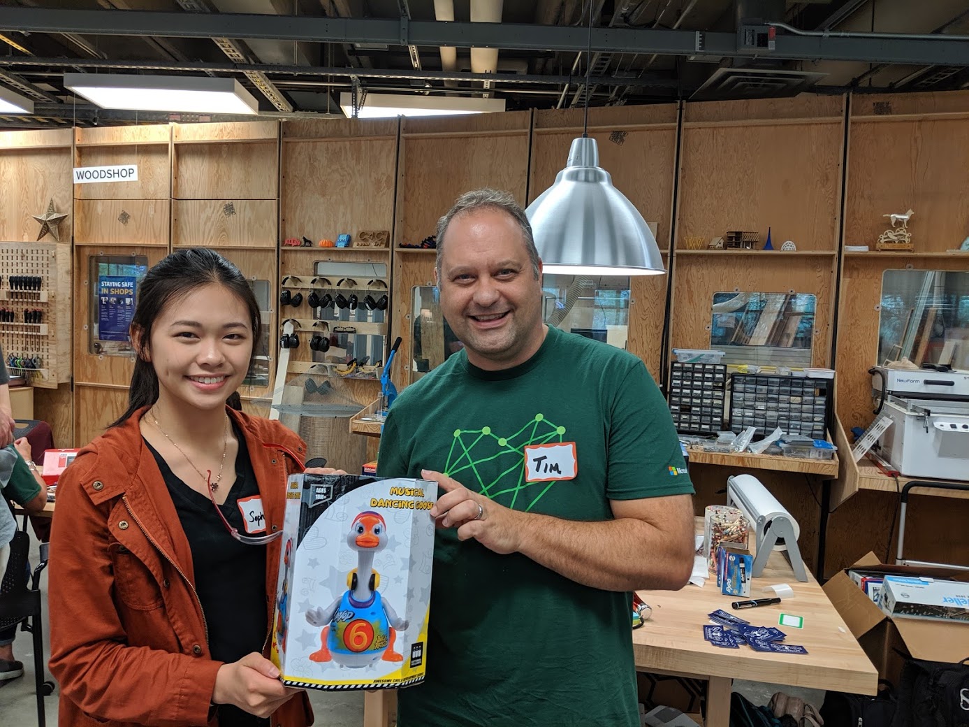 A woman wearing a orange jackets and black shirt and a man wearing a green shirt smiling while holding a toy in front of a workshop