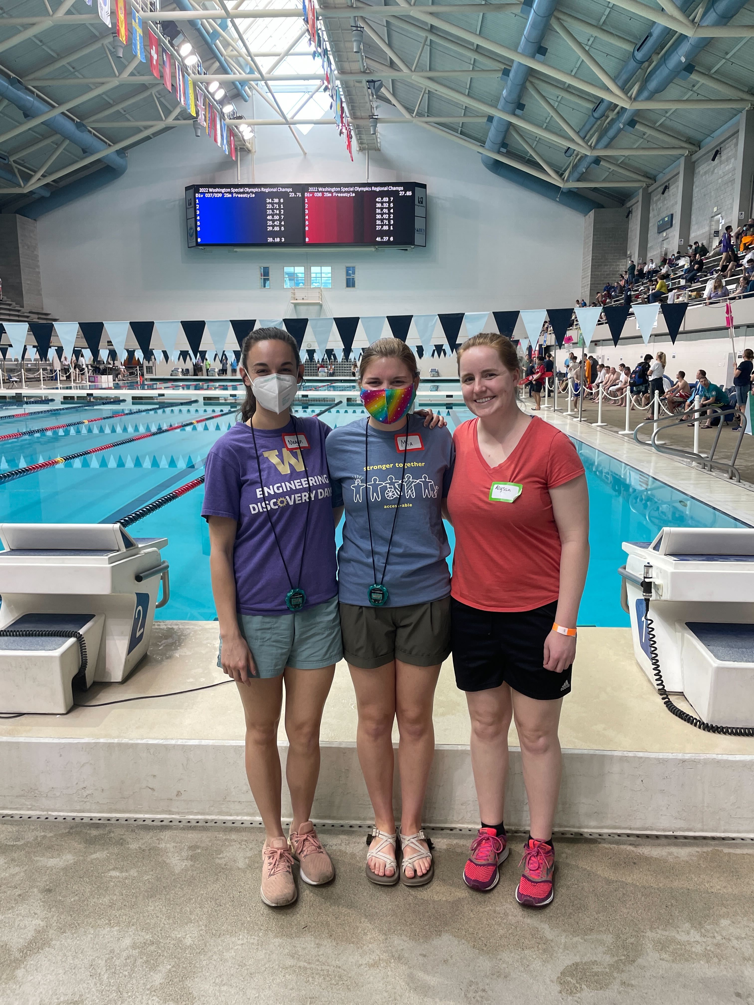 Megan, Mia, and Alyssa are standing in front of a pool with flags above wearing bright shirts.