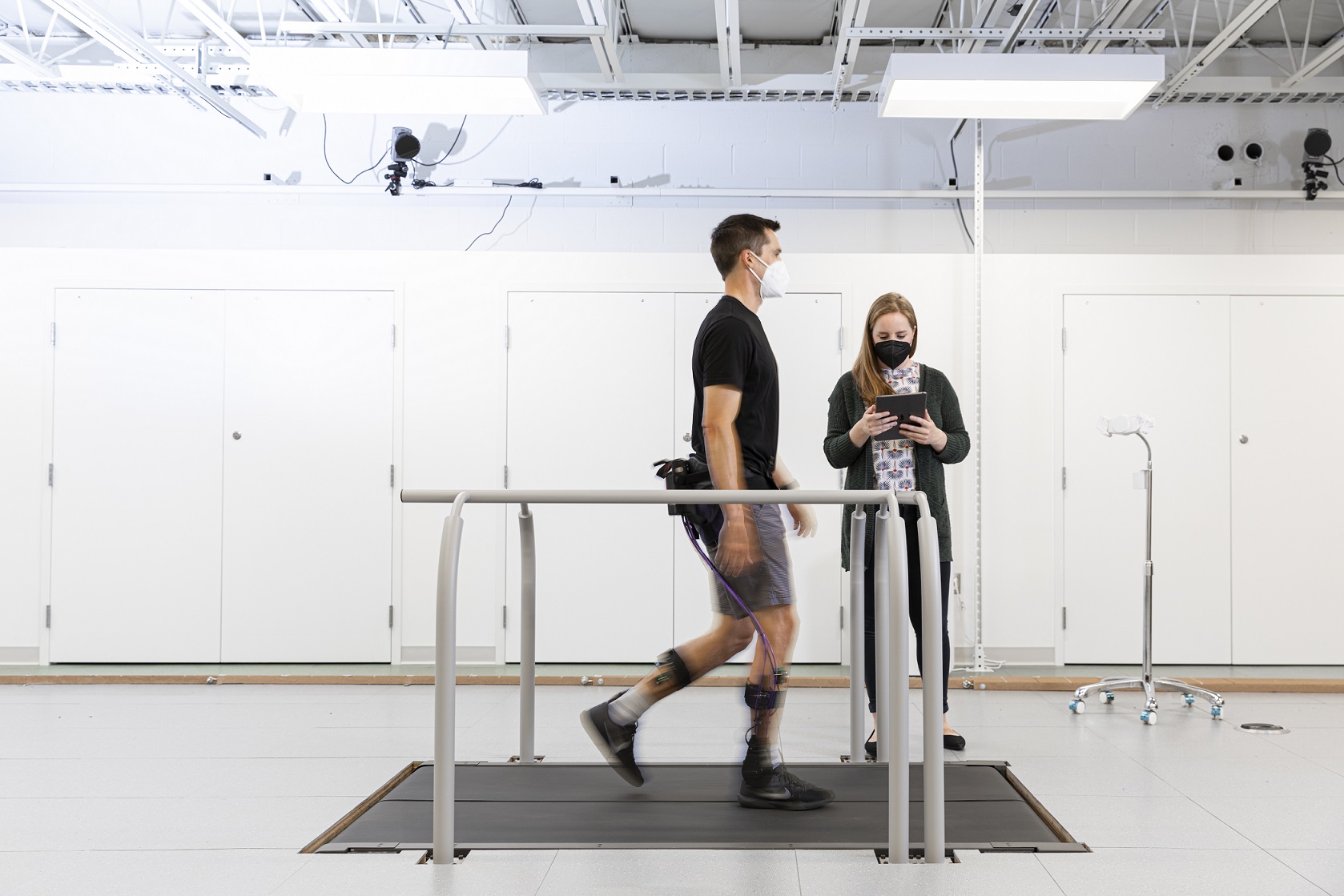 A tall man wearing an exoskeleton is walking on a treadmill in the foreground of a large white room. Alyssa is stanidng next to him looking at an ipad.