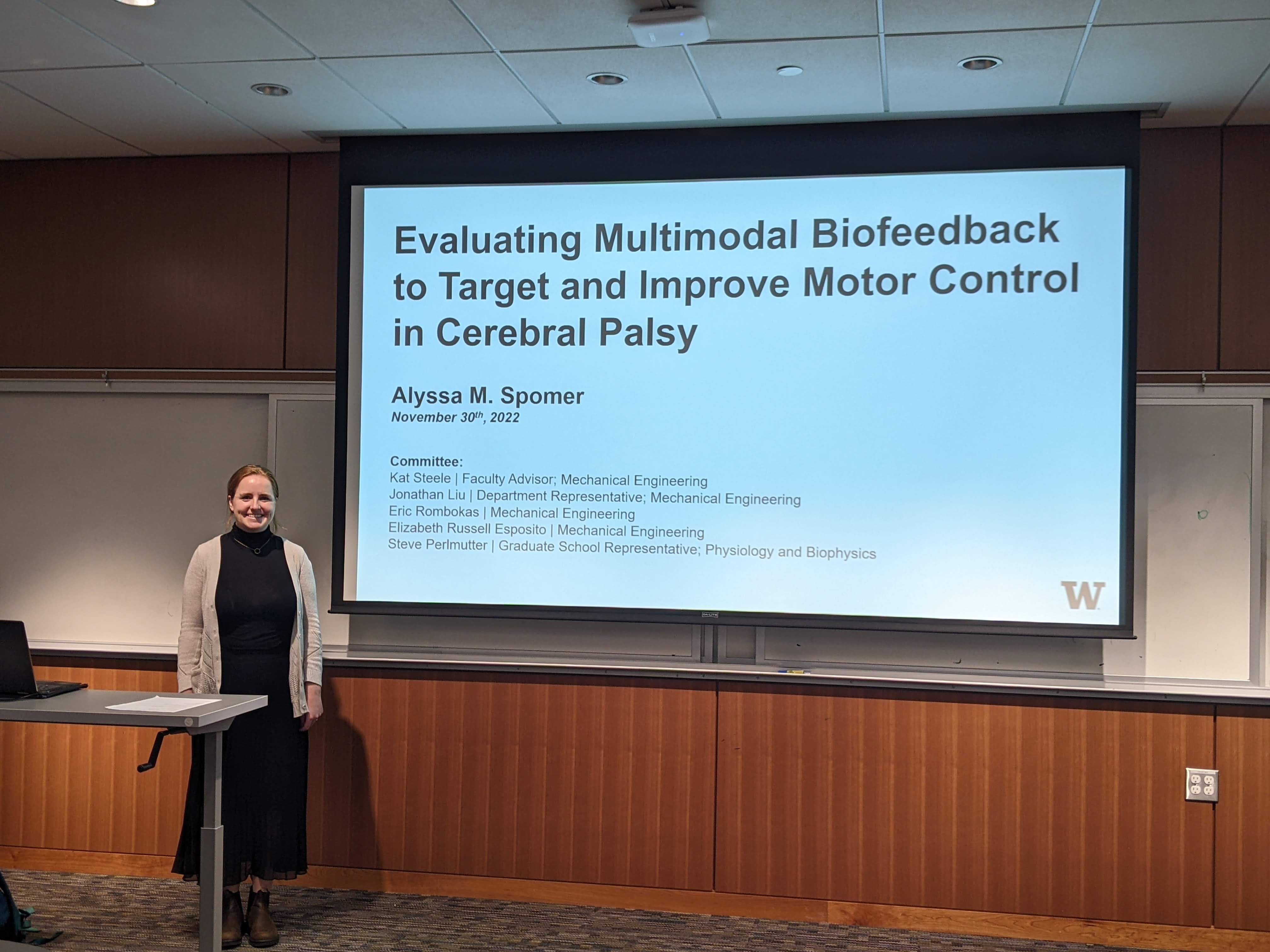 Alyssa is a white woman with red hair and is wearing a black dress and white sweater. She is smiling and standing next to a large projected screen with her dissertation title.