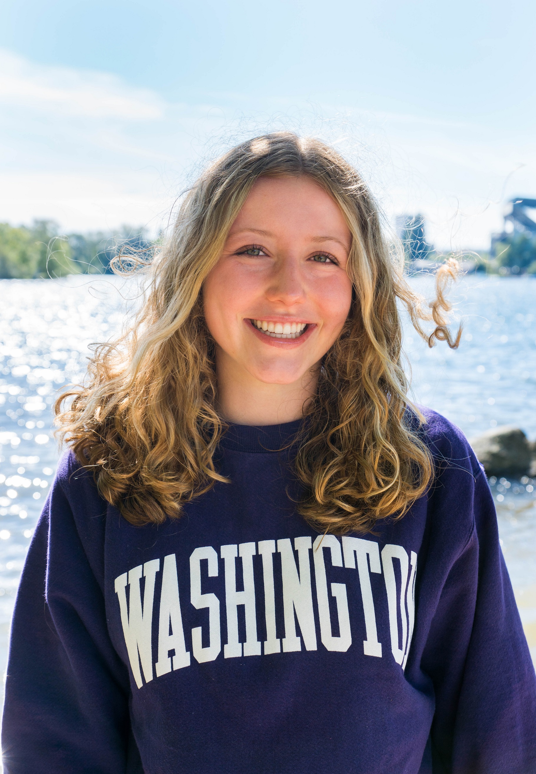 Grace has shoulder length blonde curly hair and fair skin. She is wearing a purple UW sweatshirt and is smiling in front of a body of water.