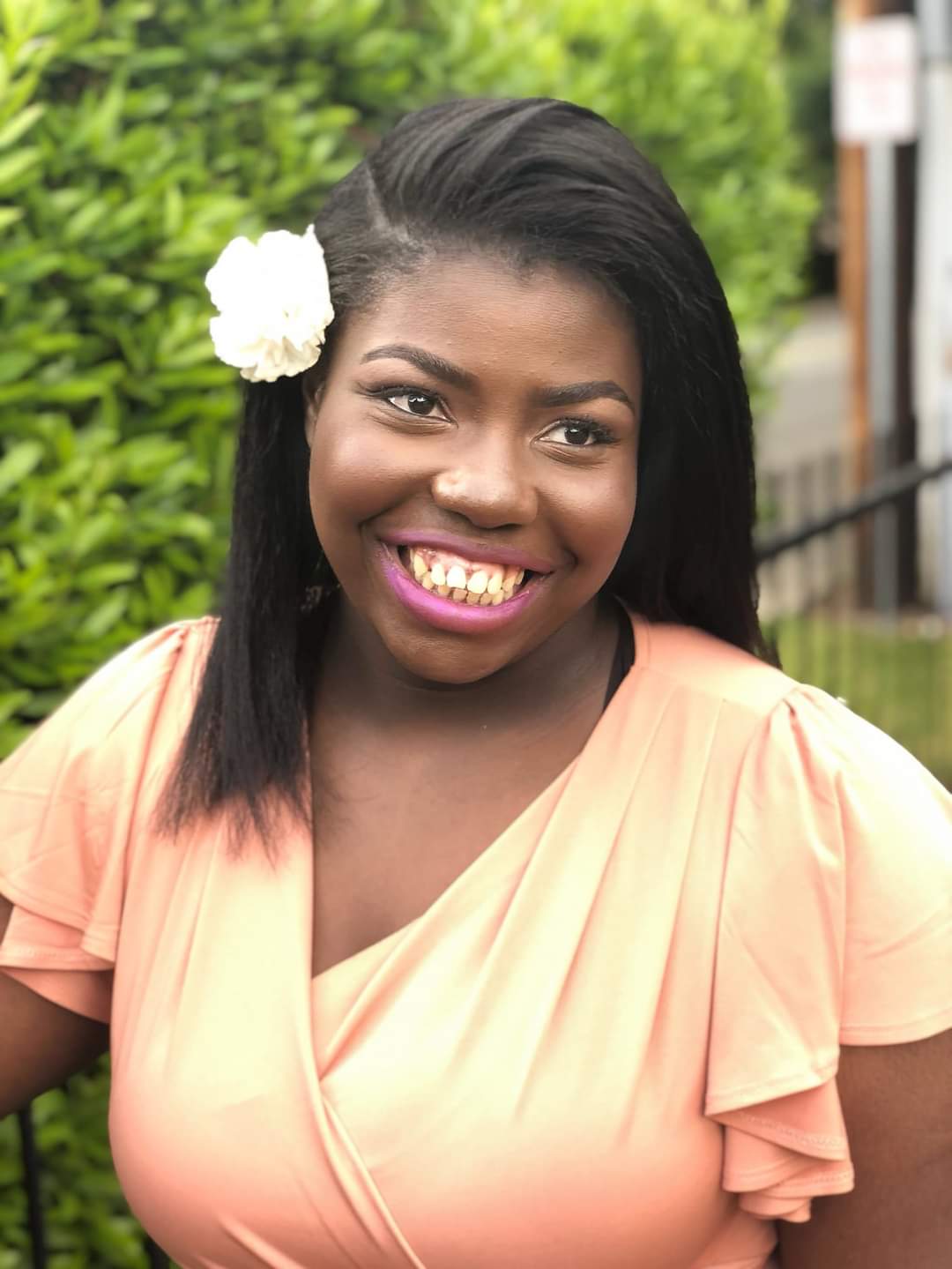 Miriam has black hair and dark skin and is wearing a white flower in her hair. She is smiling in front of greenery wearing a peach colored dress.