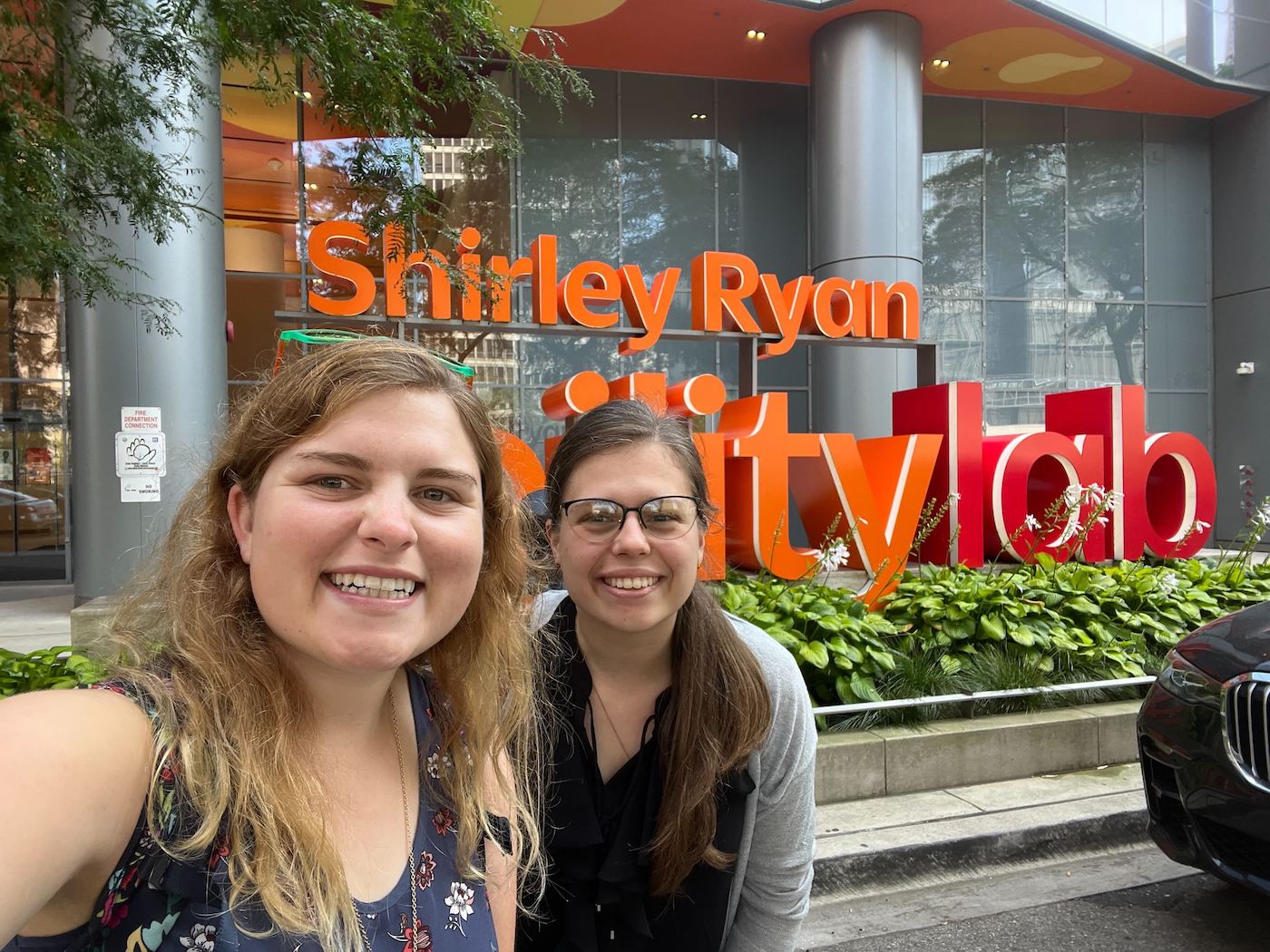 Two people smiling and taking a selfie while standing in front of The Shirley Ryan Ability Lab sign. Mia has blonde hair. Charlotte has brown hair and is wearing glasses.