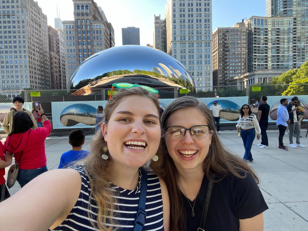 Two people smiling and taking a selfie while standing in front of The Bean in Chicago. Mia has blonde hair. Charlotte has brown hair and is wearing glasses.