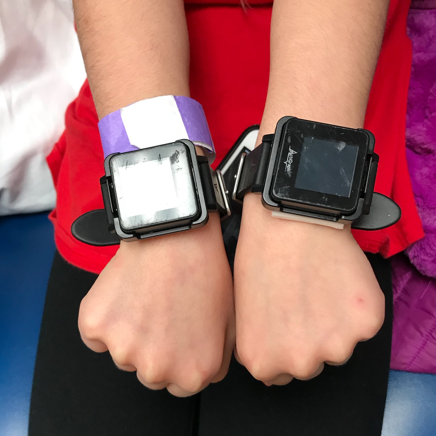 A child participant wearing an Actigraph sensor on each wrist held in front of their body. Actigraphs are IMUs that look like large black watches.