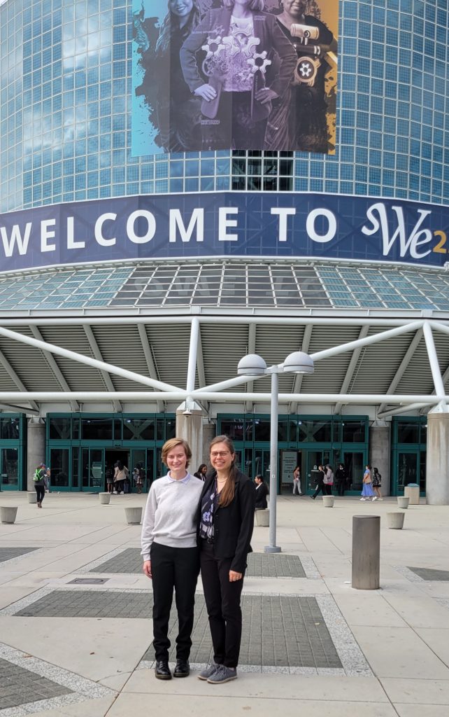 Tori and Charlotte are standing outside of a convention center with a large sign that says "Welcome to SWE 23"