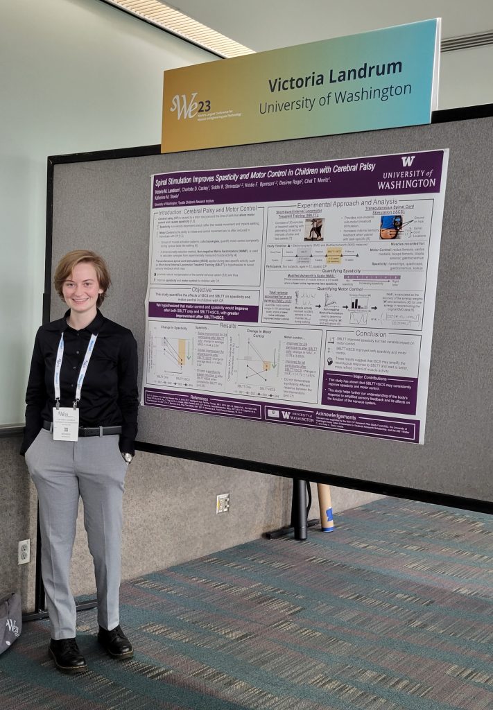 Vitoria (Tori) has short blonde hair and is wearing a black shirt and grey pant. Tori is standing in front of a poster at SWE 2023.