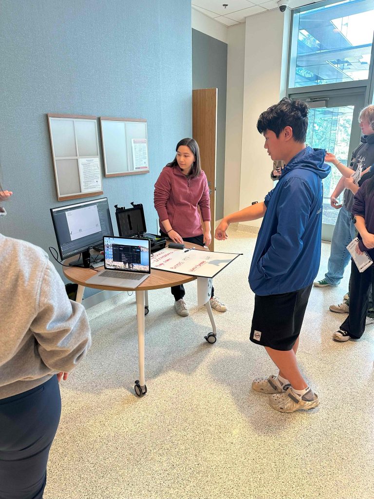 A group of people standing around a table with computers. One person is wearing an EMG sensor on their forearm