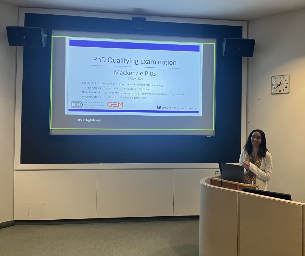 Mackenzie is standing in front of a screen displaying a slide titled "PhD Qualifying Examination"