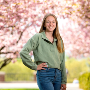 A young woman with blonde hair is wearing a green sweater and blue jeans while standing in front of a blossoming tree.