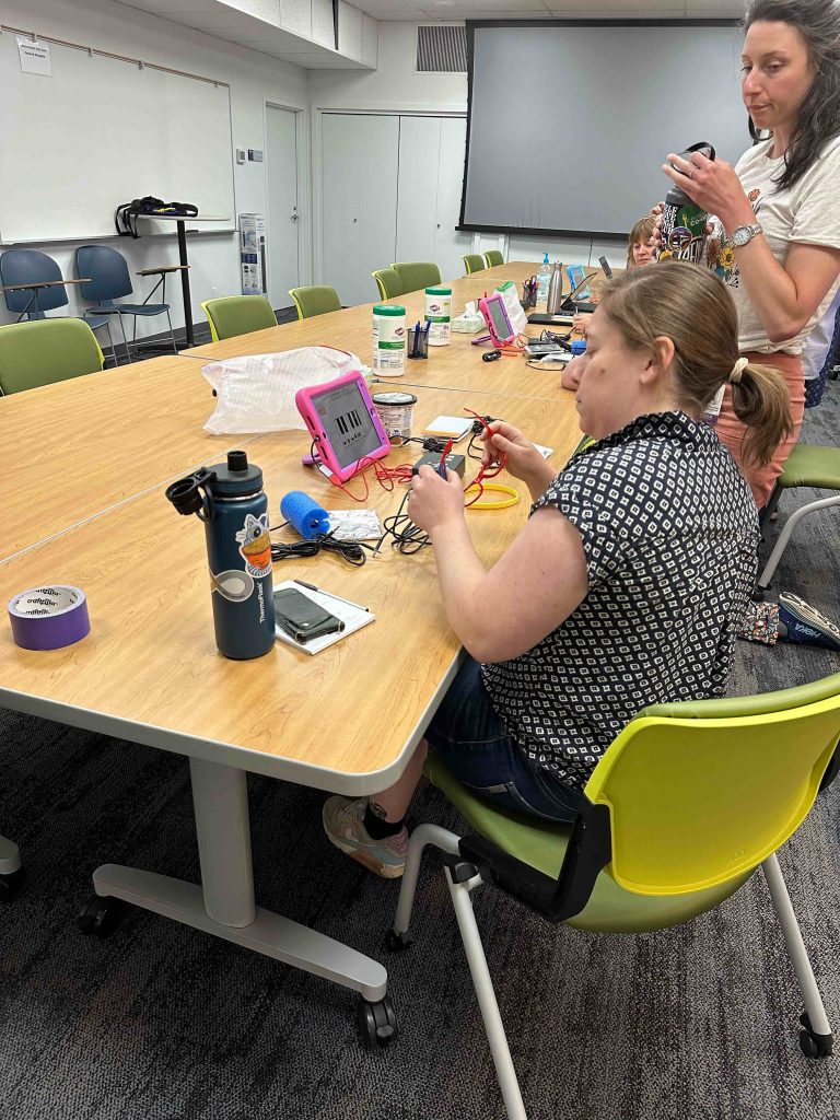 In a conference room, two women engage in hands-on work with DIY adaptive switches and an iPad.