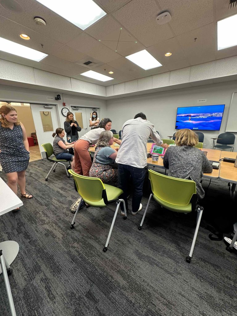 A conference room filled with people seated and standing around a large table while engaging with games and music on an iPad via DIY switches.