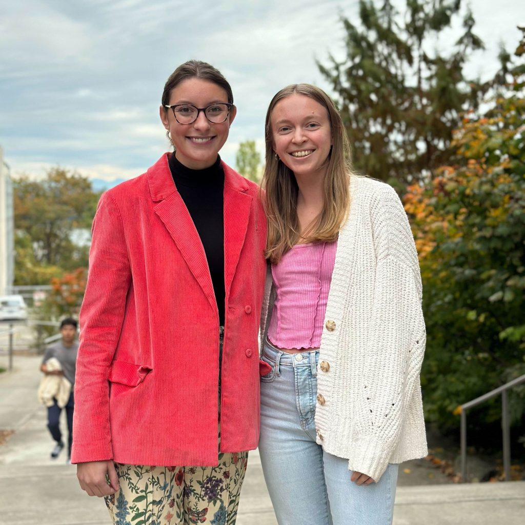 Two young women standing on a sidewalk with fall colored trees behind them. Madeleine is wear glasses and a coral colored blazer. Ally is wearing a cream cardigan sweater.