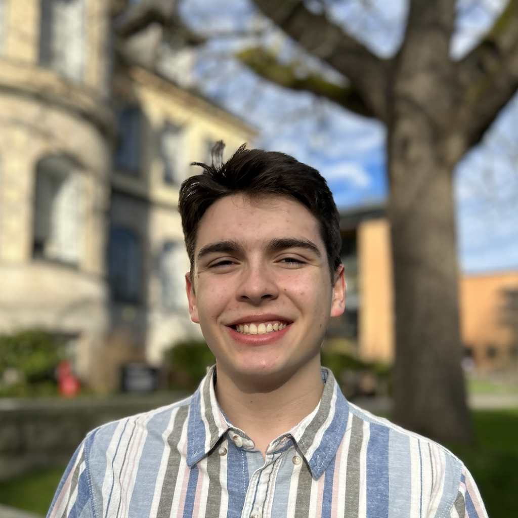 A young man smiles in front of a modern building. He has dark brown hair and is wearing a blue striped shirt.