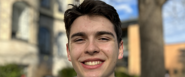 A young man smiles in front of a modern building. He has dark brown hair and is wearing a blue striped shirt.