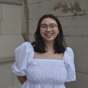 A woman with glasses smiling softly against a neutral background. She has dark brown medium length hair and is wearing a white blouse.