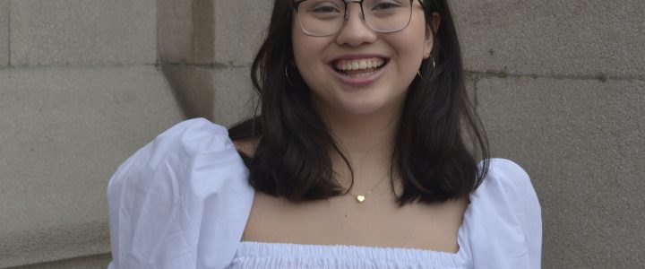 A woman with glasses smiling softly against a neutral background. She has dark brown medium length hair and is wearing a white blouse.