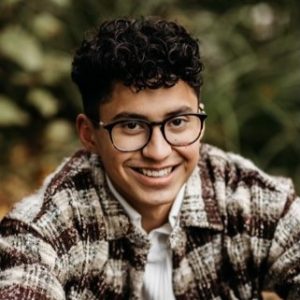 A young man smiles. He has dark brown hair, and is wearing glasses and a plaid jacket.