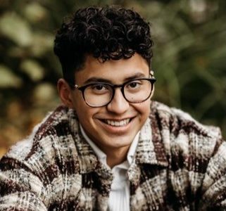 A young man smiles. He has dark brown hair, and is wearing glasses and a plaid jacket.