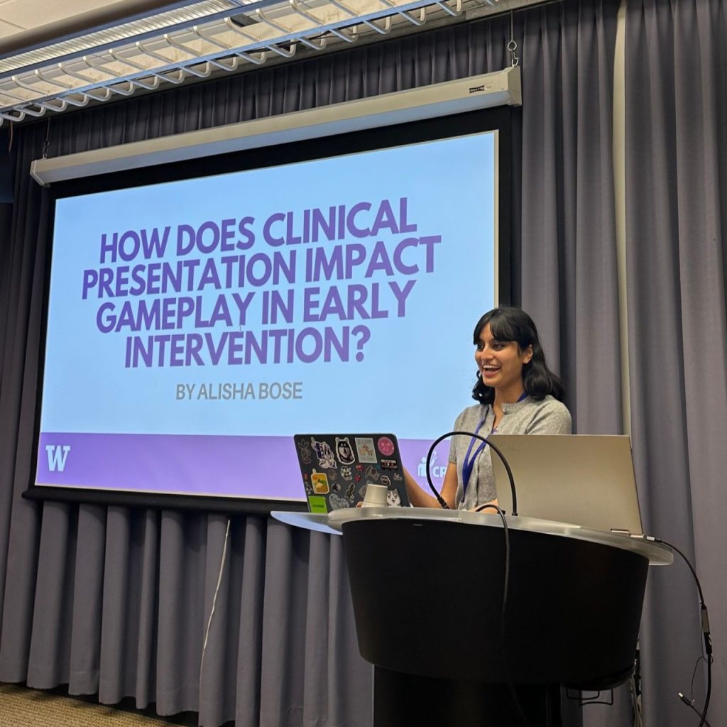 A person stands at a podium giving a presentation. Behind them, a large screen displays the title slide: “HOW DOES CLINICAL PRESENTATION IMPACT GAMEPLAY IN EARLY INTERVENTION?” by Alisha Bose.