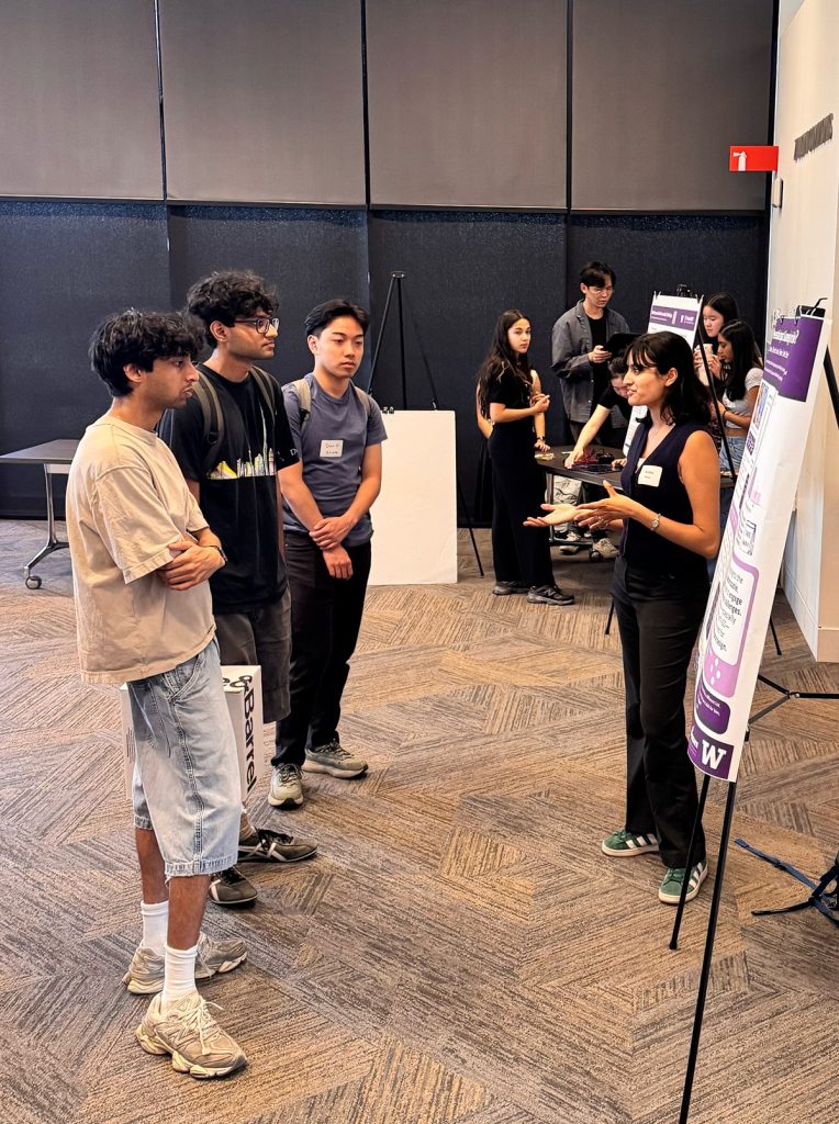 A group of people standing and conversing at an indoor research showcase. Some attendees are viewing posters or displays.