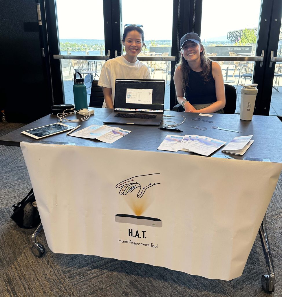 Two people seated behind a table displaying materials for the H.A.T. Hand Assessment Tool. A banner with an illustrated hand and the tool’s name is draped across the front of the table.