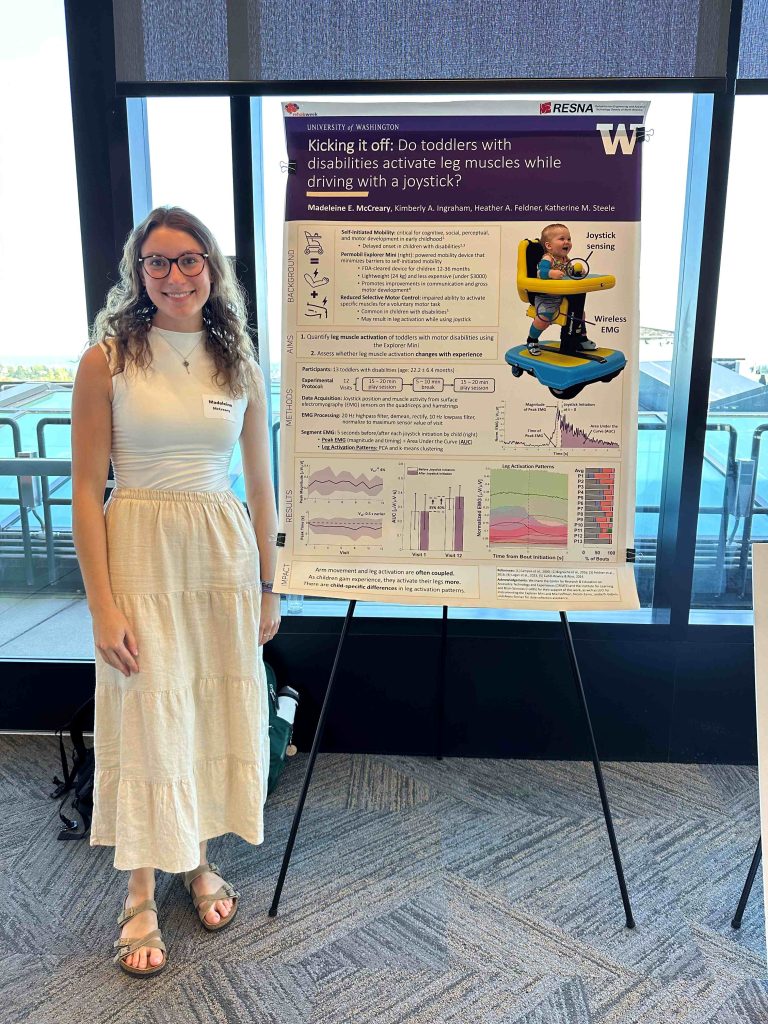 A person standing next to a scientific poster presentation titled "Kicking it off: Do toddlers with disabilities activate leg muscles when driving with a joystick?" featuring graphs, charts, and images.