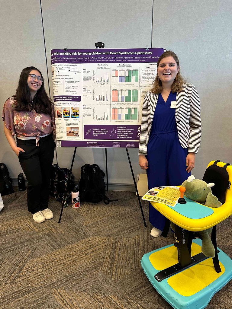 Two individuals standing in front of a research poster titled “Mobility aids to support independent play for young children with Down Syndrome” which includes graphs and charts. A small powered wheel chair with a stuffed dinosaur is set up in front of the poster.