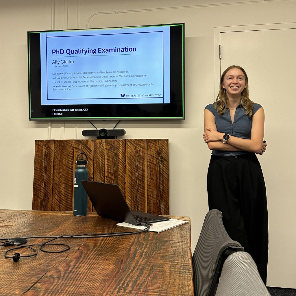 Person standing next to a large screen displaying a slide titled 'PhD Qualifying Examination' with the name Ally Clarke and University of Washington logo, in a conference room with a wooden table, laptop, and water bottle.