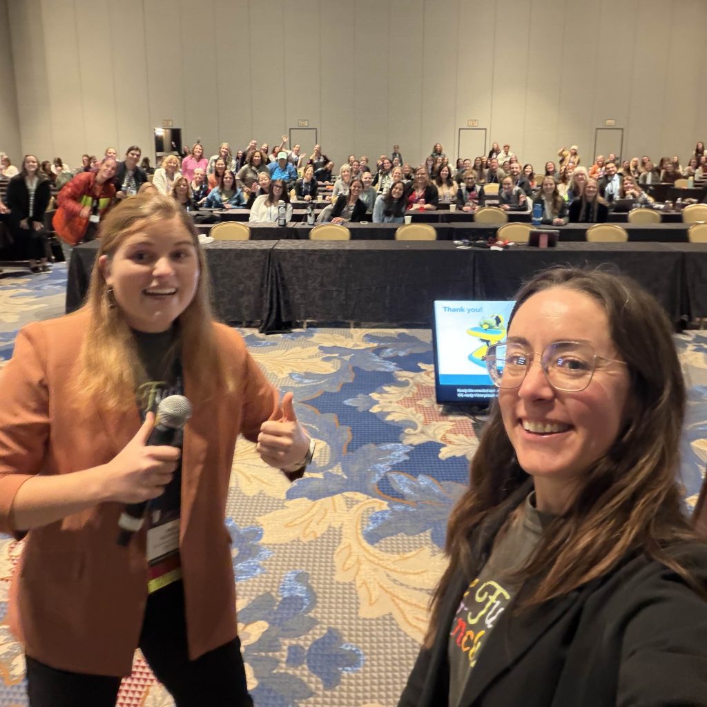 A selfie-style photo taken from the front of a large conference room, showing two presenters in the foreground and a large seated audience in the background.