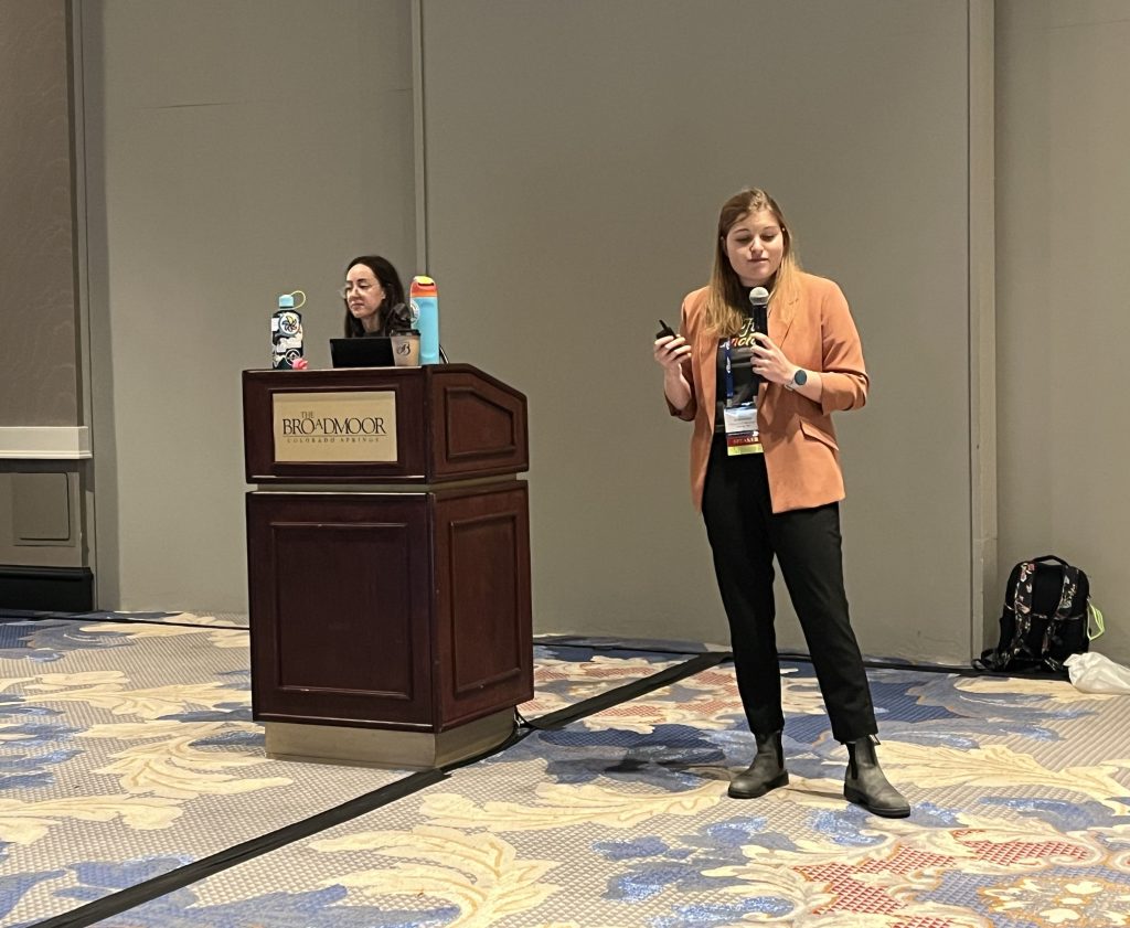 A presenter stands beside a podium with a laptop and water bottles, holding a microphone and a small device while speaking. The podium has “Broadmoor” signage, and the background shows a patterned carpet and plain wall.