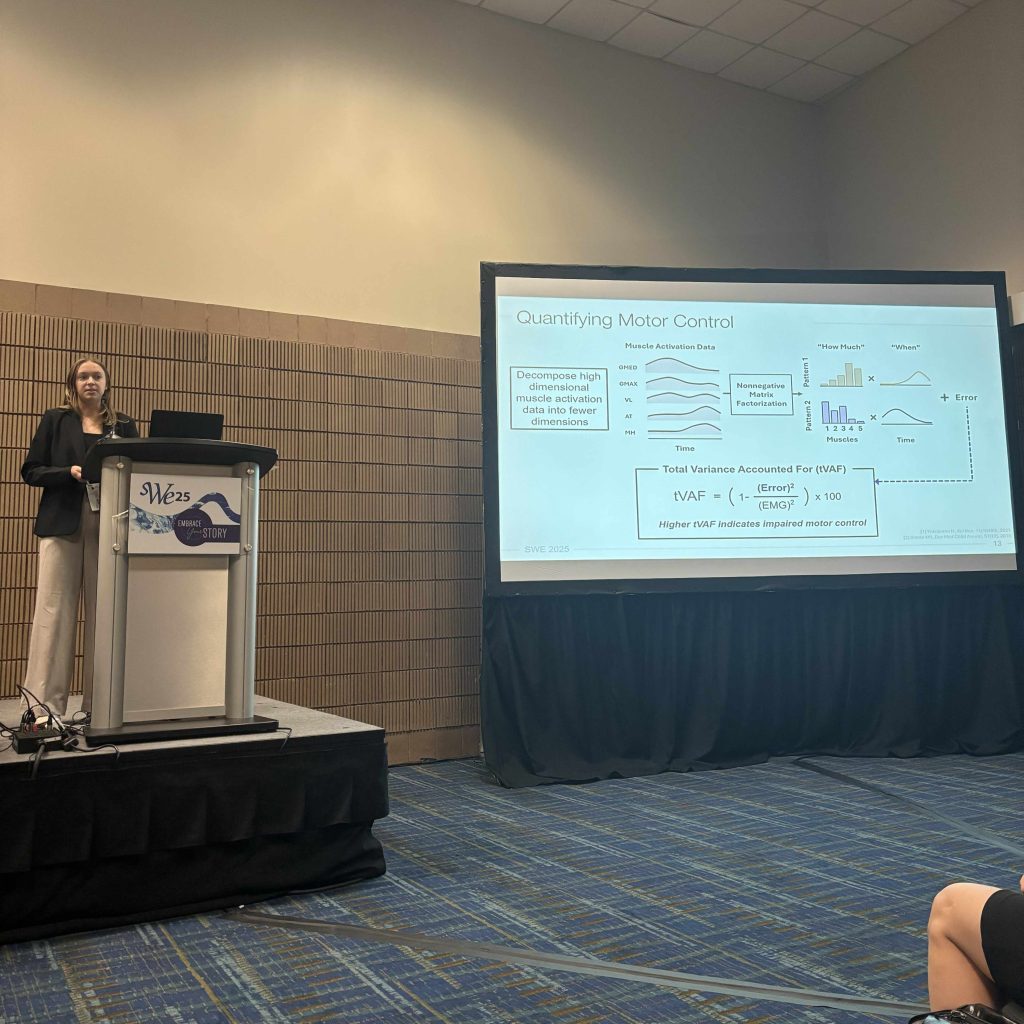 Presenter standing at a podium with a SWE25 sign, next to a large screen displaying a slide titled ‘Quantifying Motor Control’ with diagrams, graphs, and mathematical formulas. Audience chairs are visible in the foreground.