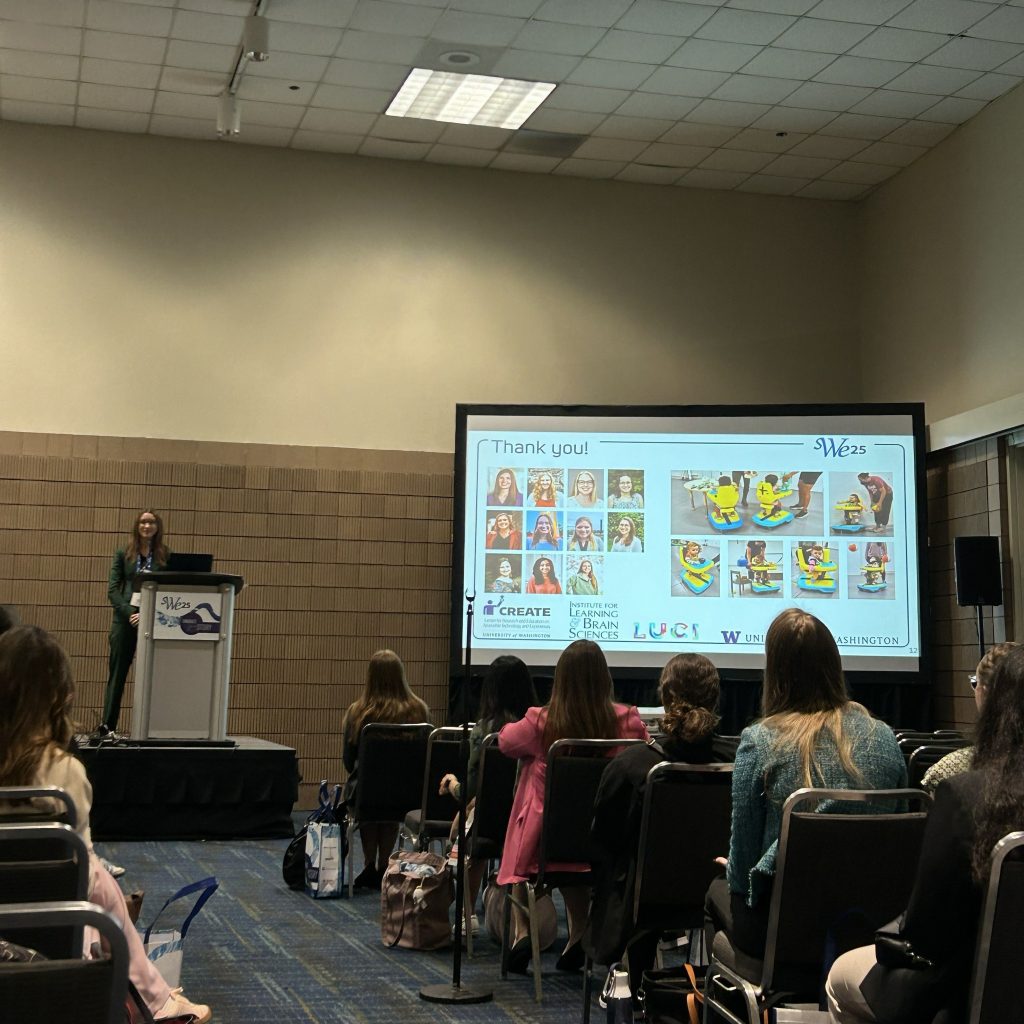 Presenter standing at a podium with a SWE25 sign, next to a large screen showing a ‘Thank You’ slide featuring photos of team members and colorful illustrations of children using assistive devices. Audience members are seated facing the screen.