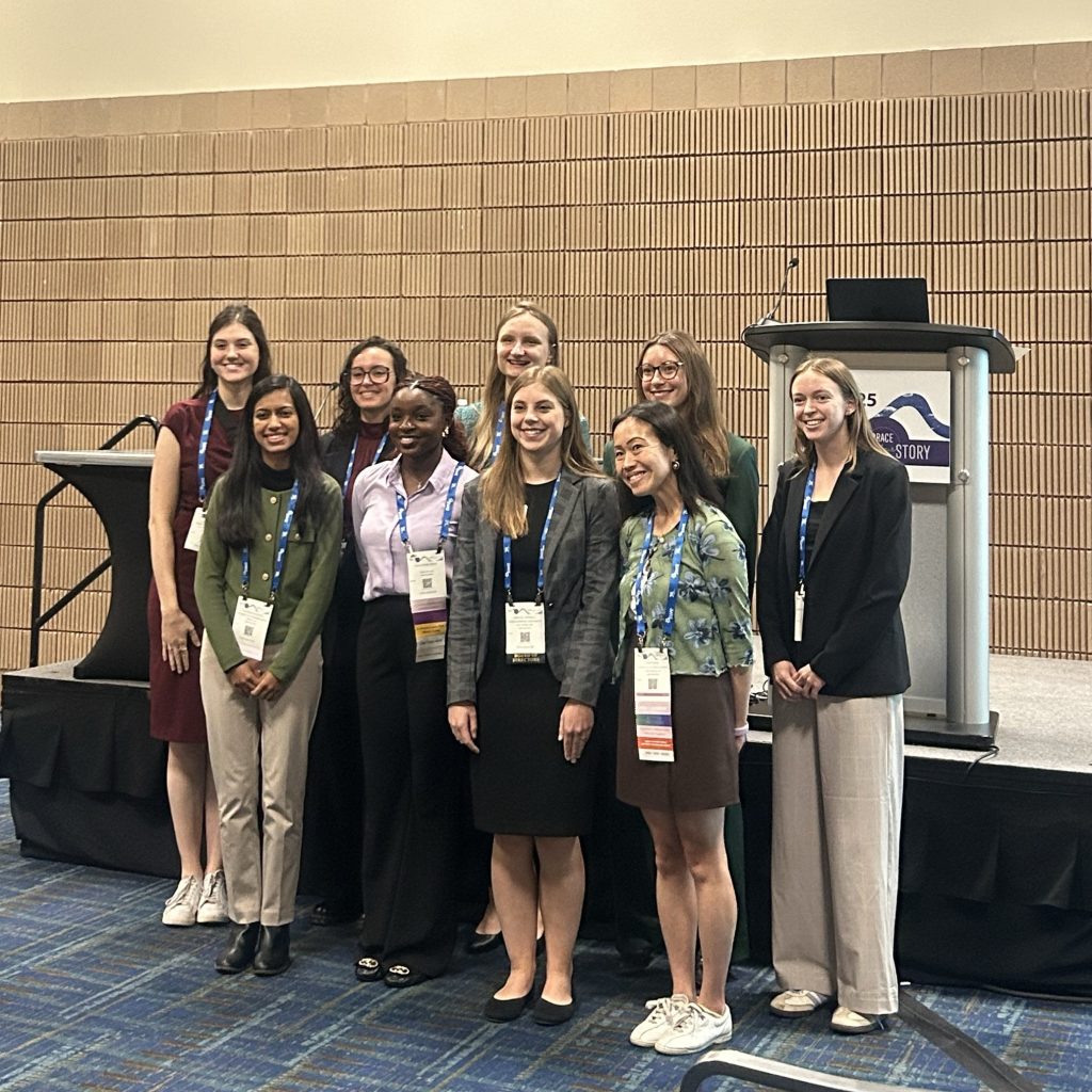 Group of individuals standing together in front of a podium with a SWE25 sign, posing for a photo in a conference room with blue patterned carpet and beige wall panels. All are wearing conference badges and lanyards.