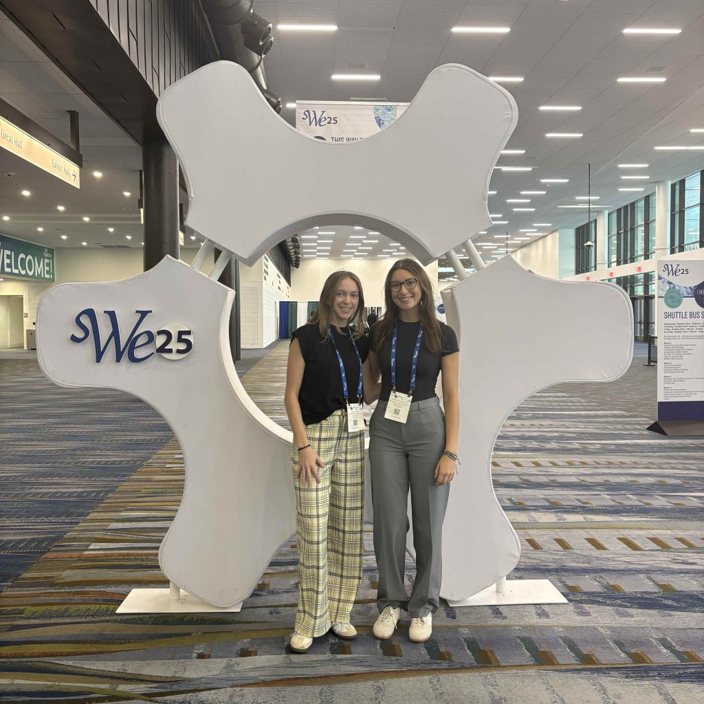 Two individuals standing in front of a large white gear-shaped display with ‘SWE25’ printed on it, inside a spacious convention center with patterned carpet and overhead lights. Both are wearing conference badges and lanyards.