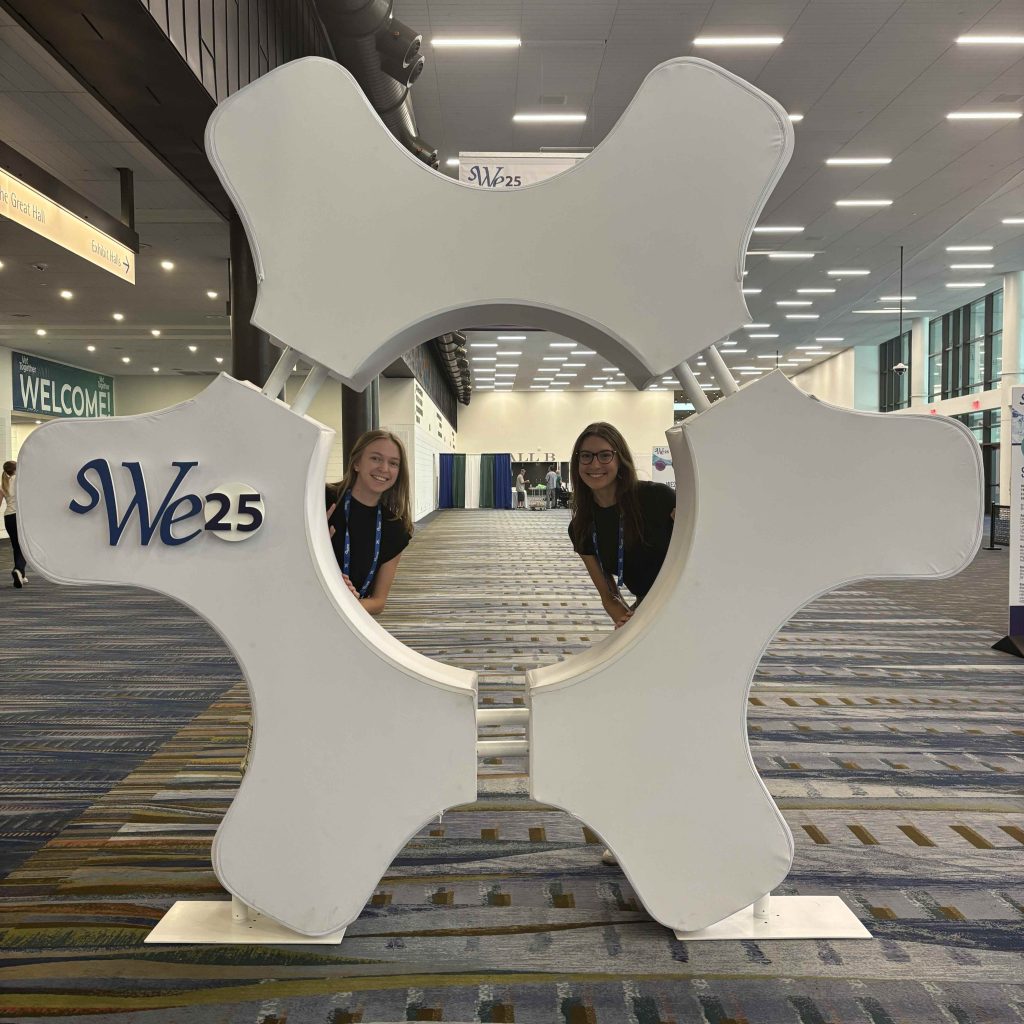 Two individuals peeking playfully through the center opening of a large white gear-shaped display with ‘SWE25’ printed on it, in a brightly lit convention center hallway.