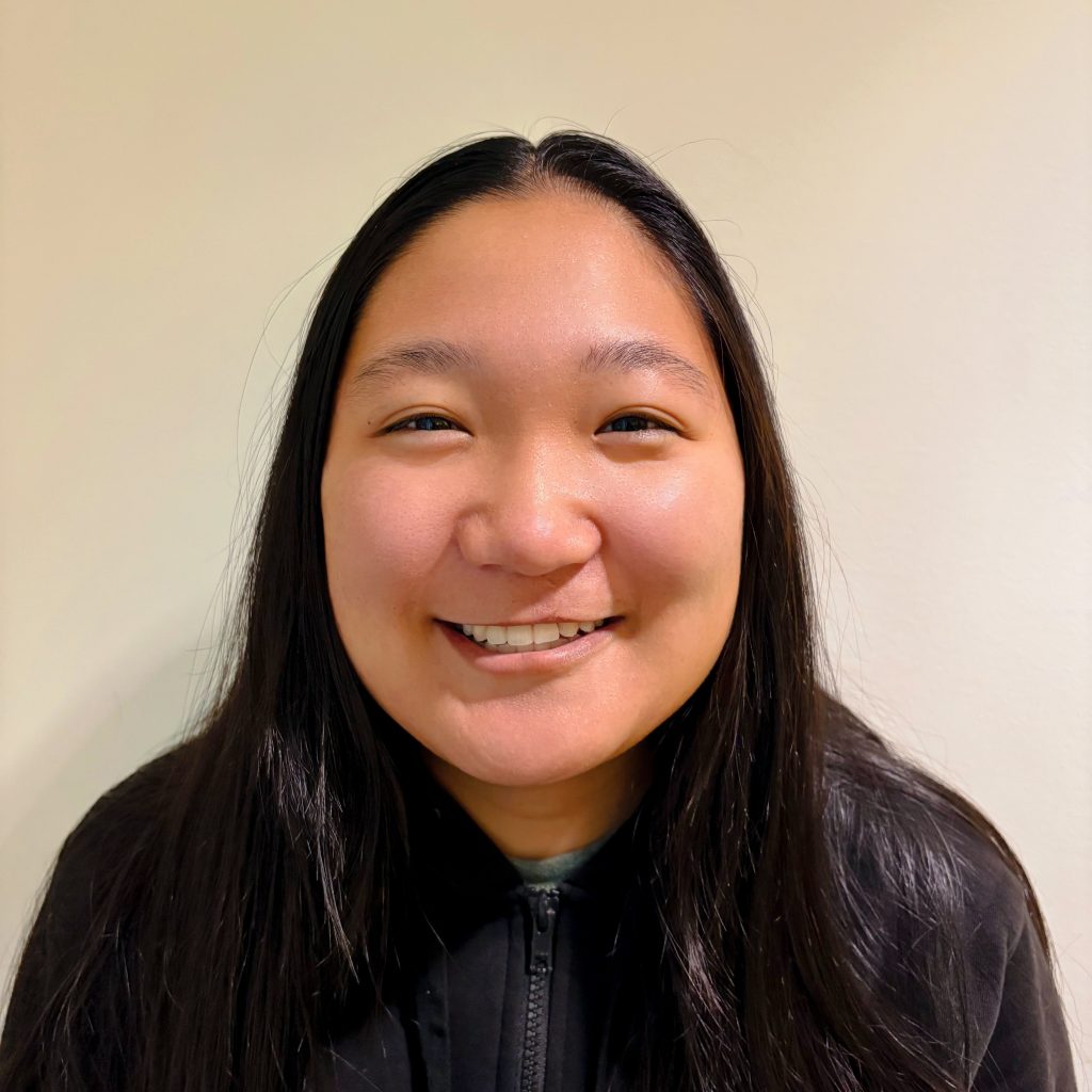Anna with long, straight dark hair, wearing a black zip-up jacket, standing against a plain light-colored background.