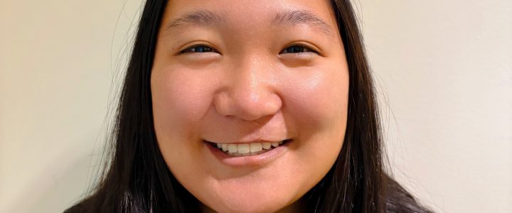 Anna with long, straight dark hair, wearing a black zip-up jacket, standing against a plain light-colored background.