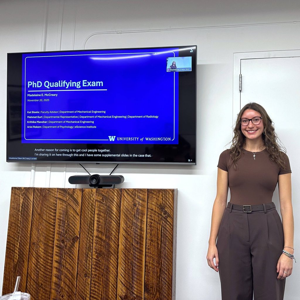 A person standing next to a large wall-mounted screen displaying a presentation slide titled ‘PhD Qualifying Exam