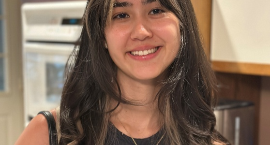 Person with long, wavy dark brown hair featuring lighter highlights, wearing a sleeveless black top and a gold pendant necklace. The individual is standing indoors in a kitchen with wooden cabinets.