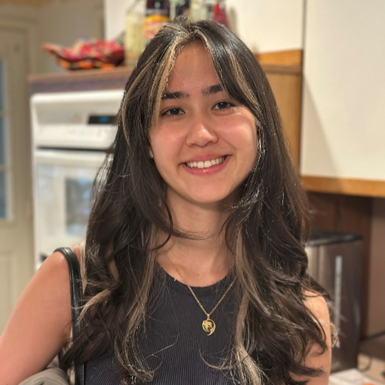 Person with long, wavy dark brown hair featuring lighter highlights, wearing a sleeveless black top and a gold pendant necklace. The individual is standing indoors in a kitchen with wooden cabinets.