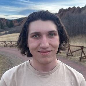 Person with medium-length wavy dark brown hair, wearing a light beige crew-neck T-shirt. Standing outdoors on a dirt path, wearing a light beige T-shirt. The background features reddish rock formations under a partly cloudy blue sky.